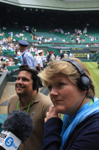 Clare Balding on centre court at Wimbledon wearing headphones with a BBC Radio 5Live microphone and Dom Joly in the background